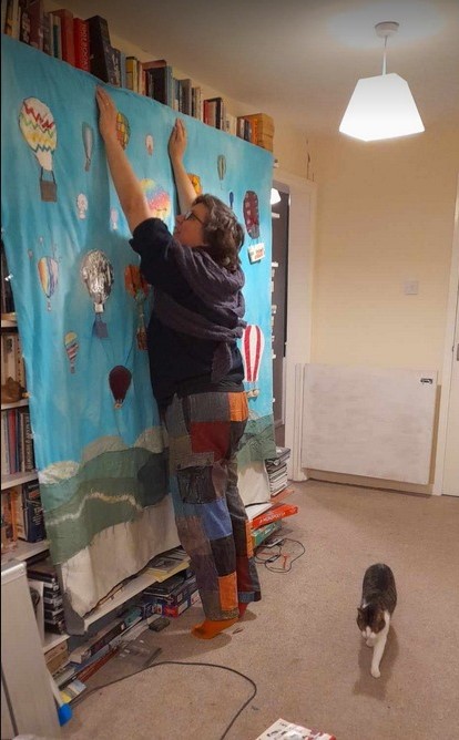The wall hanging has been somehow suspended from the top shelf of a book shelf. It starts very close to the ceiling and reaches most of the way down to the floor. A middle aged woman in patchwork trousers is stretching up next to it, trying to stop it from falling onto the floor.  A small cat walks past, unaware of the risk of balloon fall.