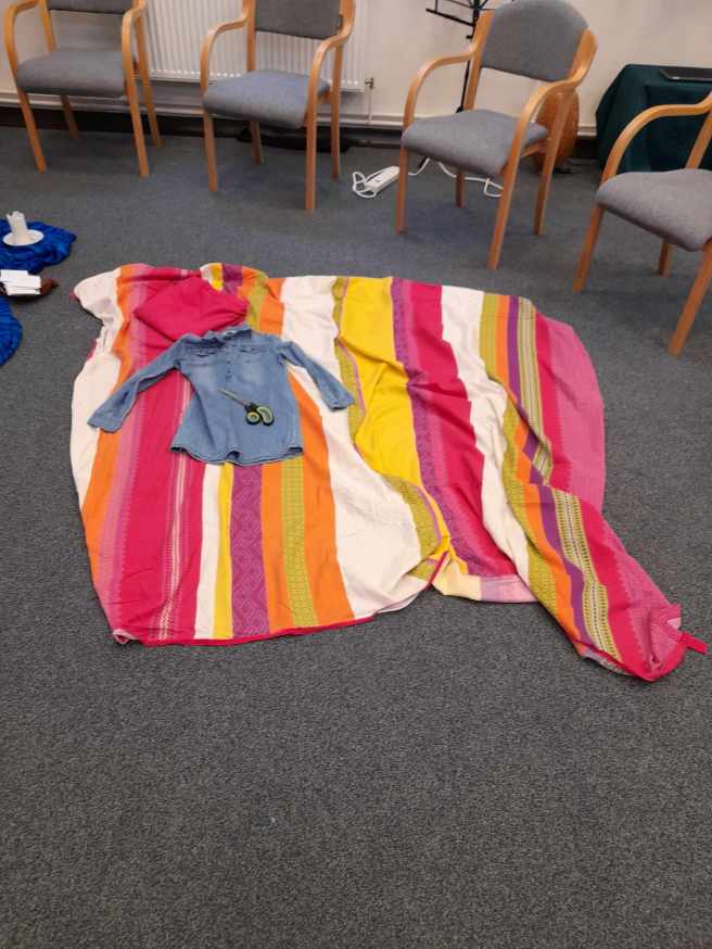 The floor of a conference room. (You can tell by the very practical grey carpet and the chair legs in view.  There's a king sized duvet cover laid out on the floor with stripes of bright colours across it. There's a denim shirt and a pair of fabric scissors lying on top of it