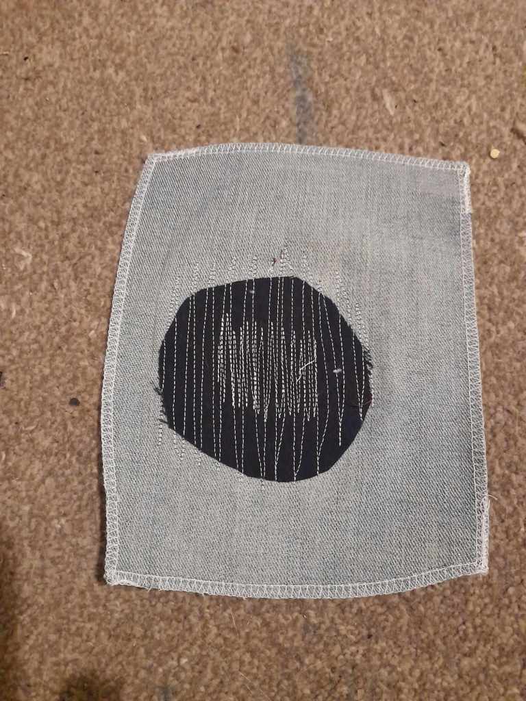 The back of the machine stitched sample. White stitching over a dark blue patch and denim. There's more stitching over the centre. The stitching goes off the edge of the patch and into the fabric around it, so the edges are held down