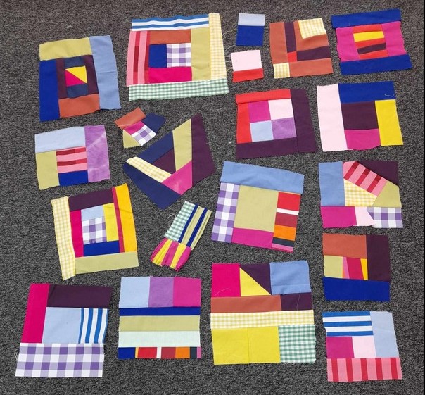 A range of patchwork blocks spread out on a dark grey carpet. There are about 16 of them, in all sorts of strong and cheerful colours. Most of the fabrics are plain, but there are some stripes and checks in their too