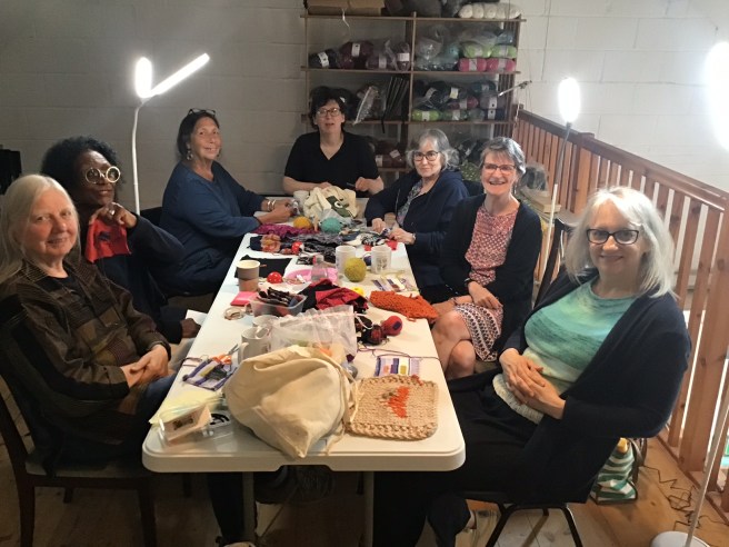 Seven women sit round a long table, which is covered with knitted samples, threads and mending paraphanalia. They look at the camera and smile in a friendly way.