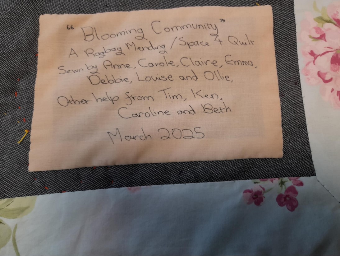 The label on the back of the quilt. The text reads: "Blooming Community" a Space 4 and Ragbag Mending Quilt sewn by Anne, Carole, Claire, Emma, Debbie, Louise and Ollie. Other help from Tim, Ken, Caroline and Beth. March, 2025